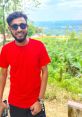 Smiling man in a red t-shirt and sunglasses, surrounded by lush greenery, enjoying a scenic outdoor view.