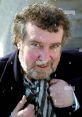 Paul Bradley smiling while adjusting his scarf, showcasing his curly hair and distinctive beard in a casual outdoor setting.