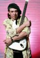 Musician embracing a white guitar, wearing a colorful shirt and a black beret against a soft pink background.