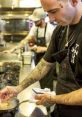 Chef Douglas Rodrigues de Castro preparing a dish in a busy kitchen, showcasing his culinary skills and tattoos.