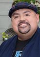 Gabriel Iglesias smiling, wearing a plaid hat and dark jacket, with greenery in the background, showcasing his cheerful personality.