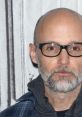 Moby, the American musician and producer, with a gray beard and glasses, poses against a simple background.