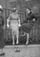 Two stylish women laughing in 1950s fashion, sitting on a fence with high heels nearby, capturing a joyful moment.