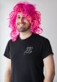 Man with a vibrant pink curly wig, wearing a black t-shirt featuring a stylish "n" logo, exuding a fun and playful vibe.