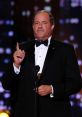 Chris Berman in a tuxedo delivering a speech at an awards ceremony, showcasing his signature charismatic style.