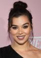 Hailee Steinfeld smiling confidently at a red carpet event, showcasing her beauty and stylish look against a pink backdrop.