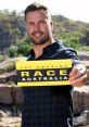 Host of The Amazing Race Australia holding the iconic yellow and black race sign against a scenic backdrop.