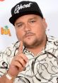 Charlie Sloth poses confidently, wearing a black cap with "Ghost" and a stylish patterned shirt, showcasing his signature style.