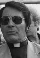 Jim Jones wearing sunglasses, a clergy collar, and a serious expression, symbolizing his controversial leadership.