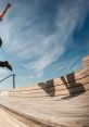 Skateboarder performing an impressive jump over a wooden ramp under a blue sky with wispy clouds.