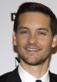 Tobey Maguire smiles at a public event, showcasing his stylish black suit and charming demeanor.