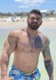 Muscular man with tattoos at the beach, wearing colorful swim trunks, showcasing confidence and summer vibes.