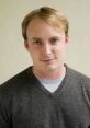 Young man with light brown hair, wearing a gray sweater, smiling against a neutral background. Professional portrait style.